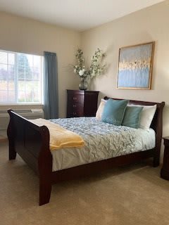 A cozy bedroom with elegant wooden furniture, a bed adorned with soft pillows and a blanket, decorated with a painting above and natural light streaming through the window.