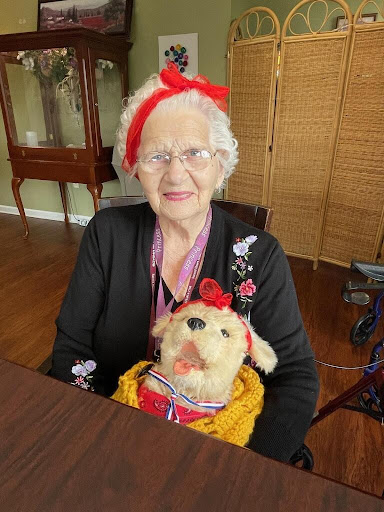 An elderly woman cheerfully poses with her dog plushie, dressed in festive colors.