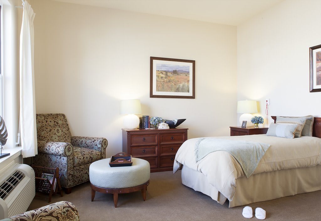 A cozy bedroom with an armchair, ottoman, dresser, and a neatly made bed.