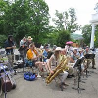 A band performs outdoors with various musicians playing instruments like saxophones in a wooded area.