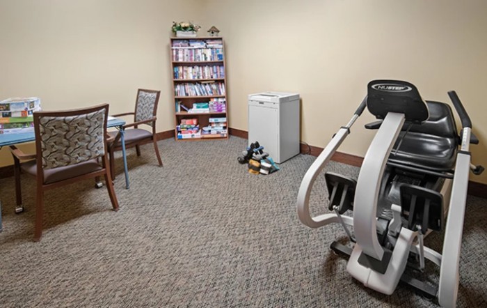 A small office space with an exercise machine, bookshelf, chairs around a table, and a mini fridge.