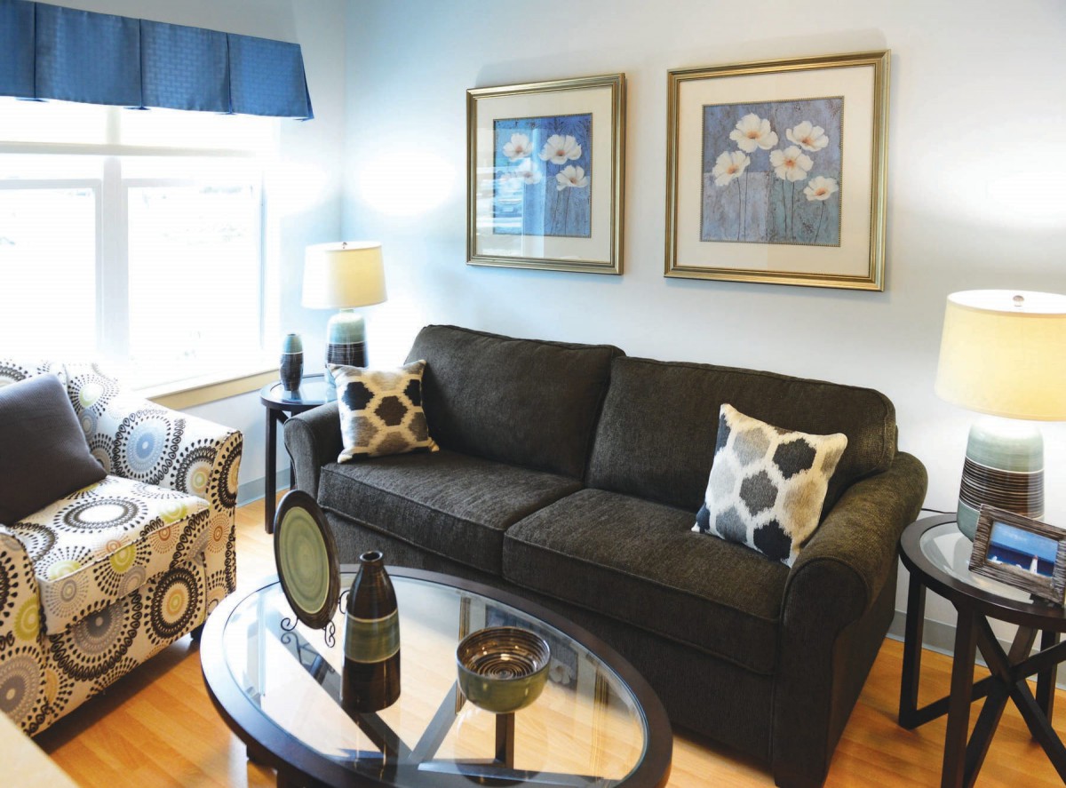 A cozy living room with a dark sofa, patterned armchair, and glass coffee table, accented by framed artwork and decorative pillows.