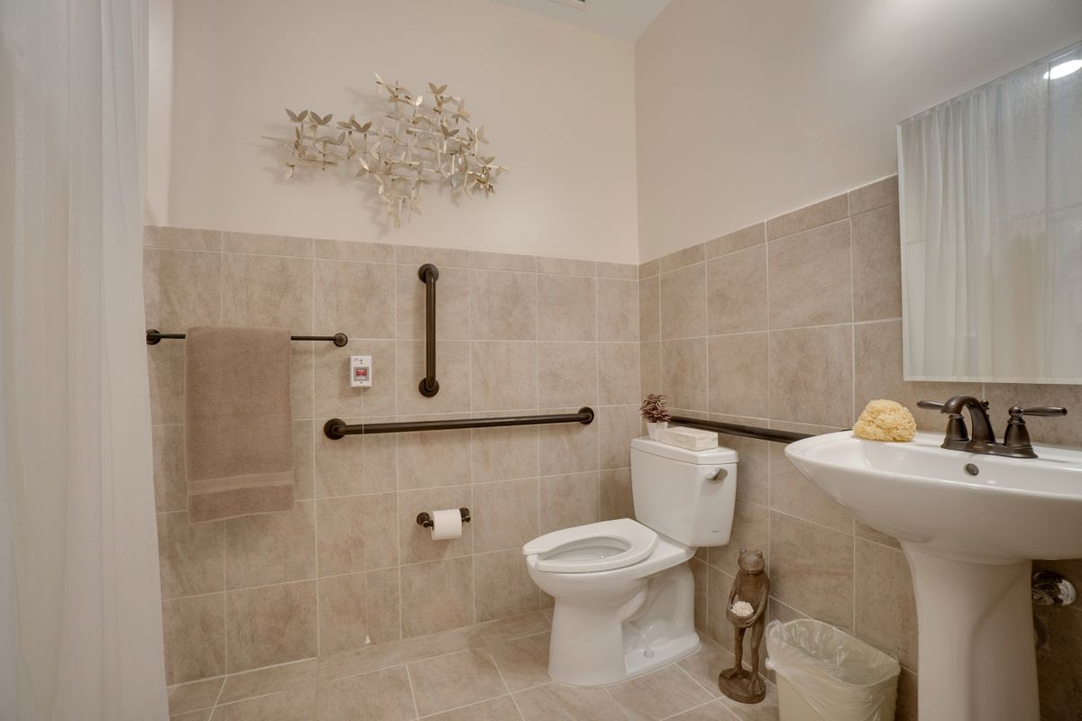A beige-tiled bathroom featuring a toilet, sink, towel rack, wall art, and accessibility grab bars.