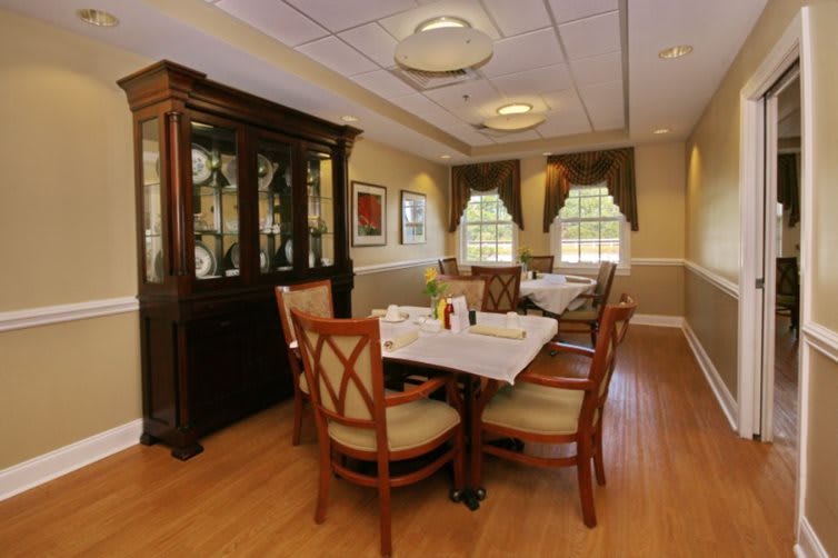 A cozy dining room with a neatly set table and chairs, accompanied by a wooden cabinet and sunlit windows.