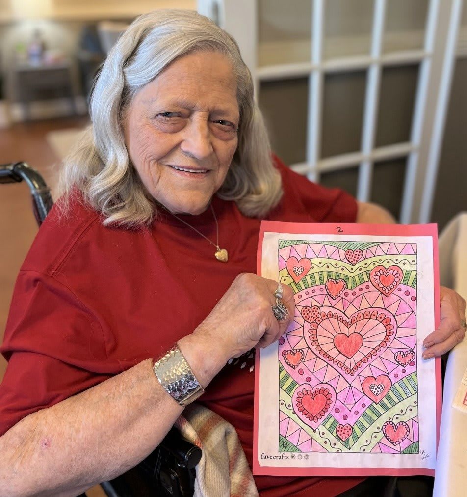 An elderly woman smiles while holding her colorful artwork featuring a heart design.