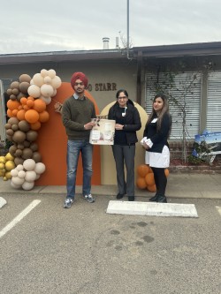 A group of three individuals presenting an award or certificate at a festive outdoor event adorned with colorful balloons.