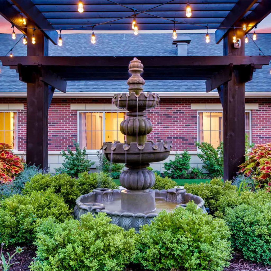 A beautifully landscaped garden with a central fountain surrounded by greenery, under a pergola with string lights.