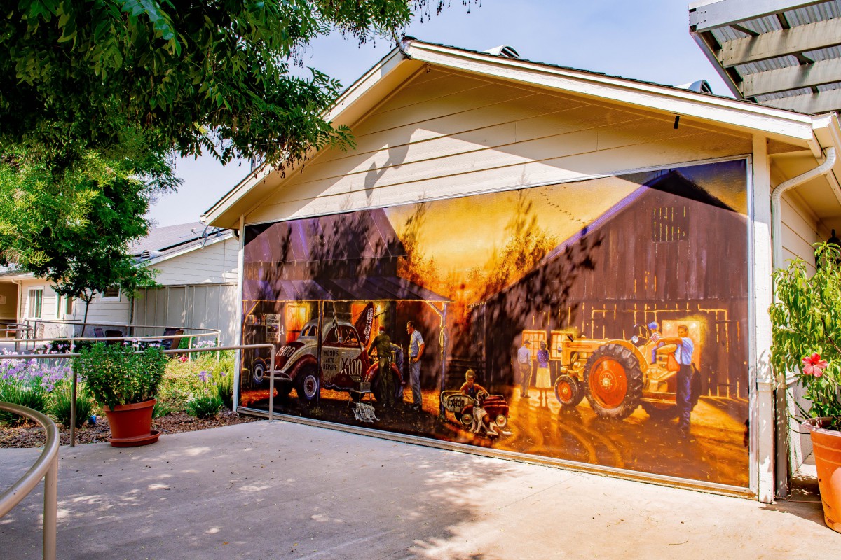 A vibrant mural featuring firetrucks covers the side of a garage next to a driveway, surrounded by trees.