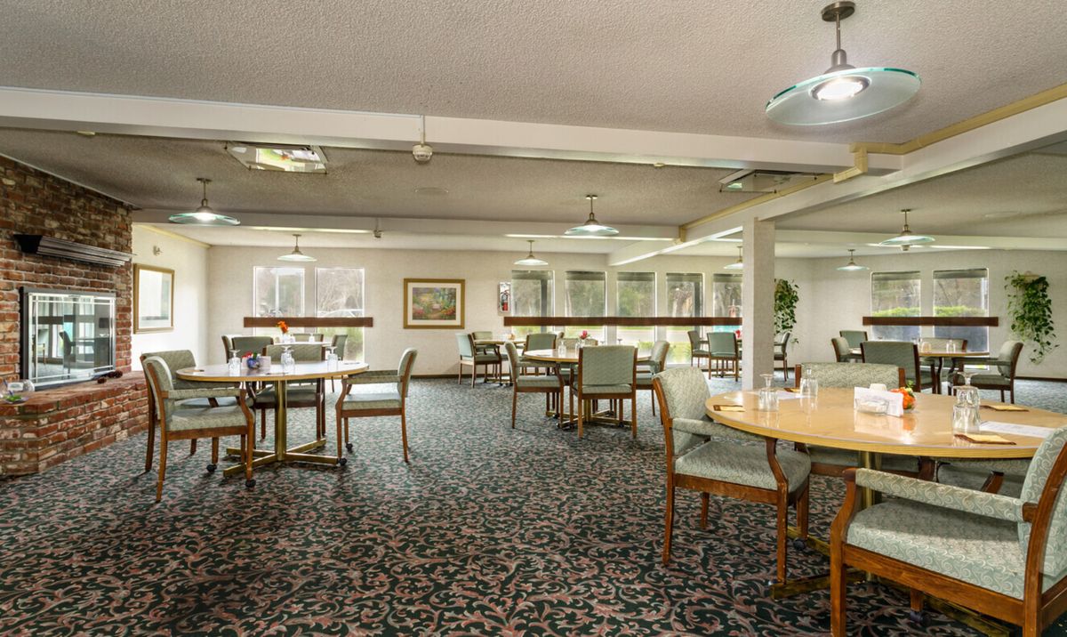 A spacious dining room with multiple tables and chairs, featuring patterned carpet and ceiling lights.
