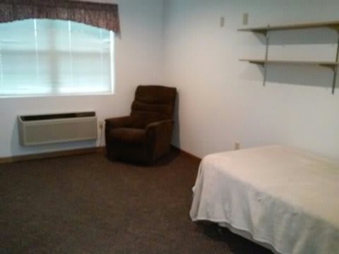 A simple room with a bed, chair, and shelves.