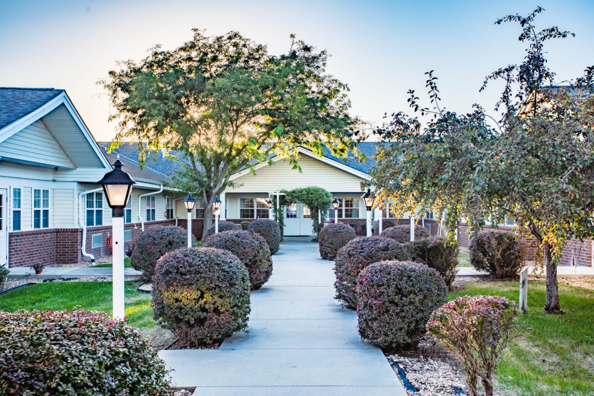 A serene outdoor view featuring a landscaped pathway leading to a building, lined with neatly trimmed bushes and trees.