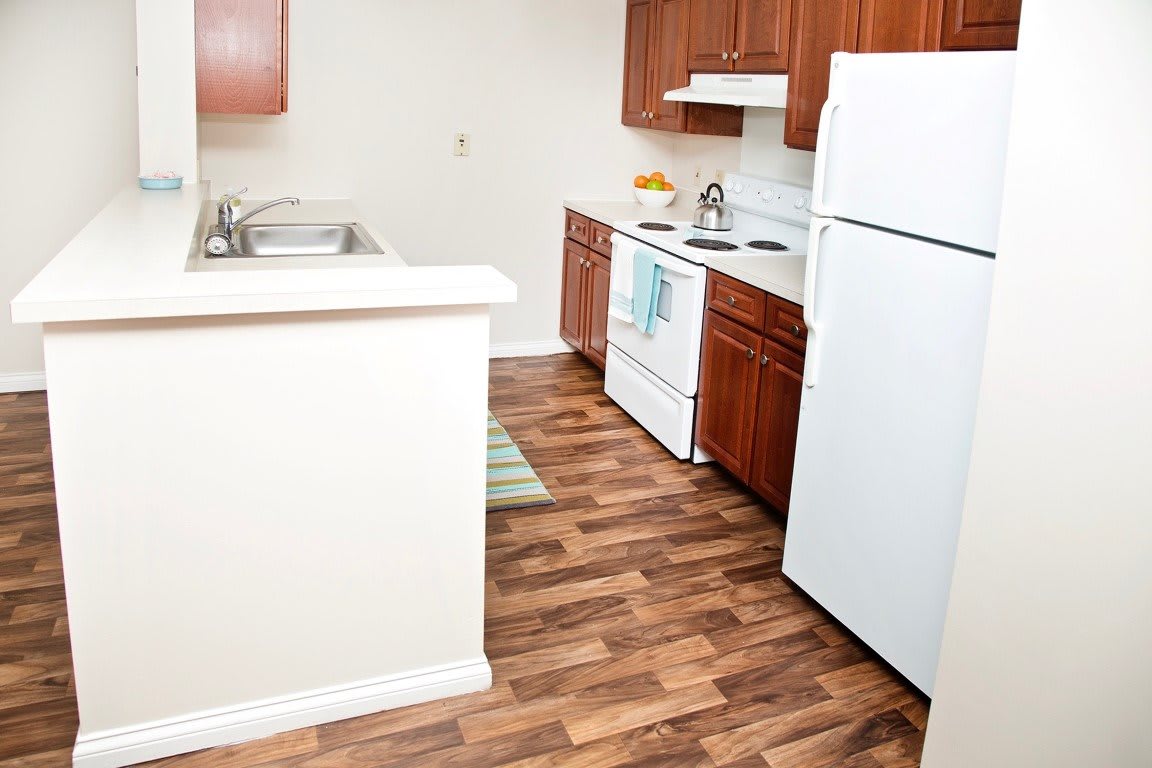 A compact apartment kitchen with wooden cabinets, a refrigerator, stove, and hardwood flooring.