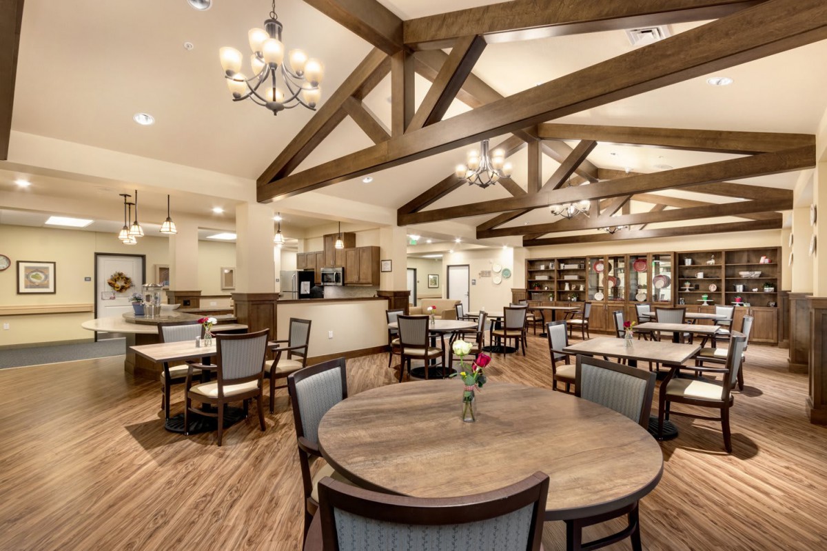 A warmly lit dining area with wooden beams, chandeliers, and neatly arranged tables and chairs.