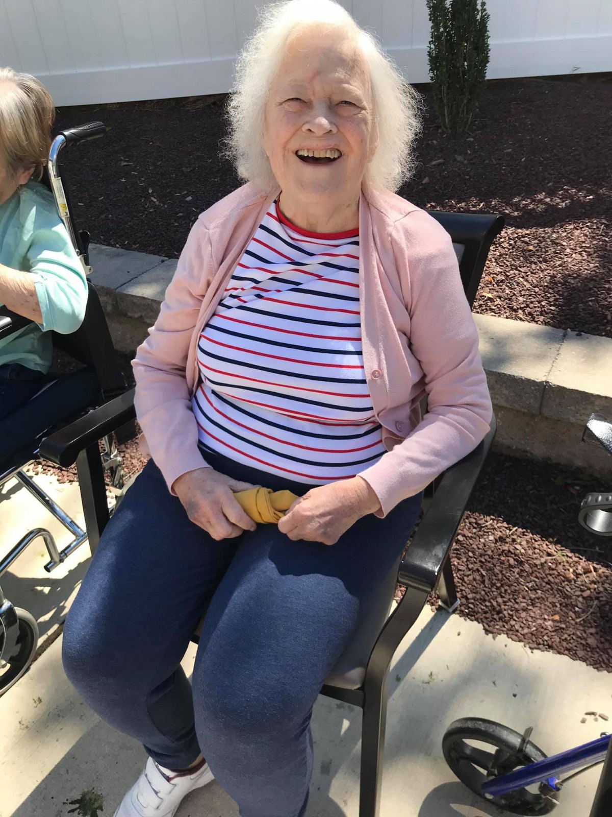 An elderly woman sitting outdoors on a sunny day, wearing a striped shirt and smiling.