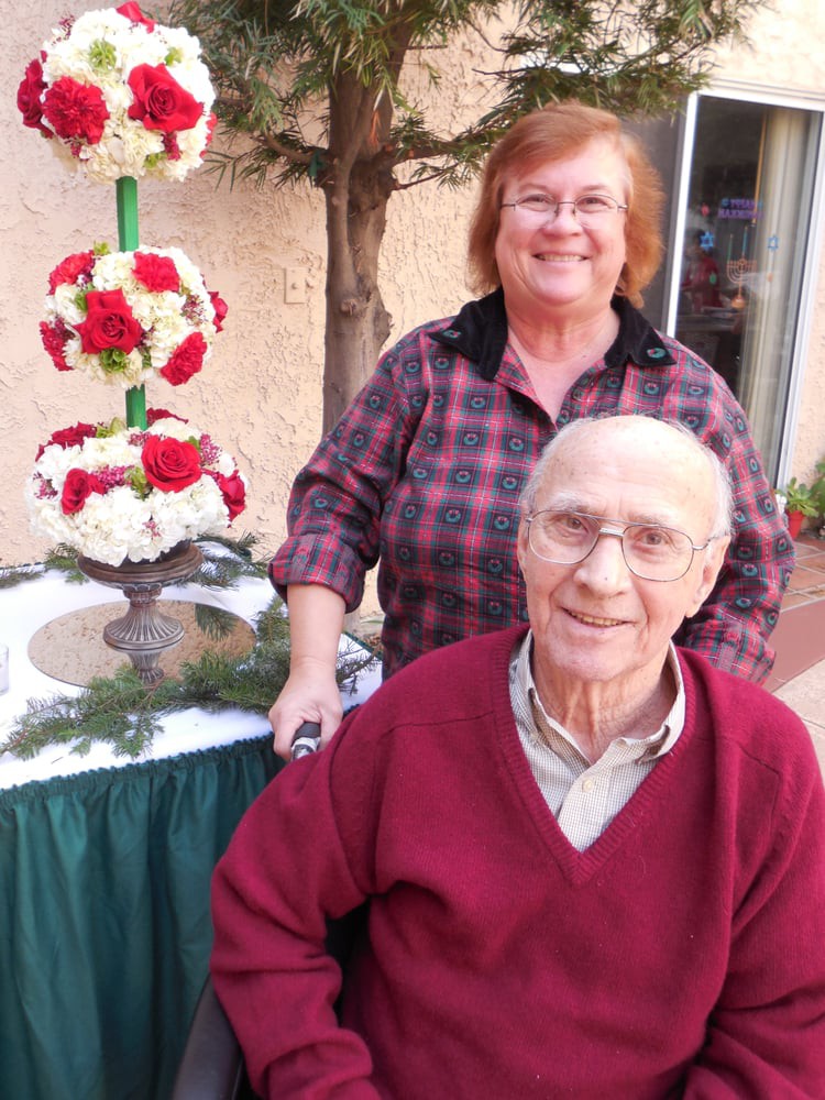 An elderly couple smiling outdoors with a floral arrangement in the background.