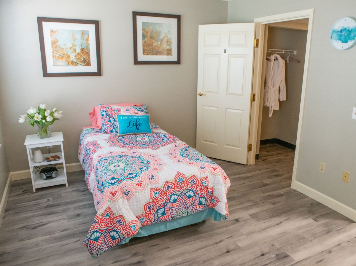 A cozy bedroom with a single bed featuring colorful bedding and decorative cushions, complemented by artwork on the walls and a wooden floor.