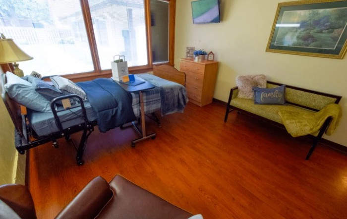 A cozy bedroom with a single bed, wooden flooring, and a comfortable chair next to large windows.