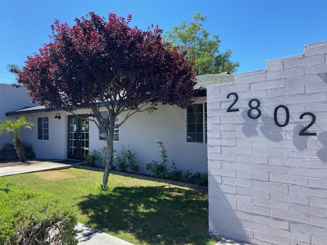 A suburban house with a tree and well-maintained lawn, featuring the number 2801 on a brick wall.