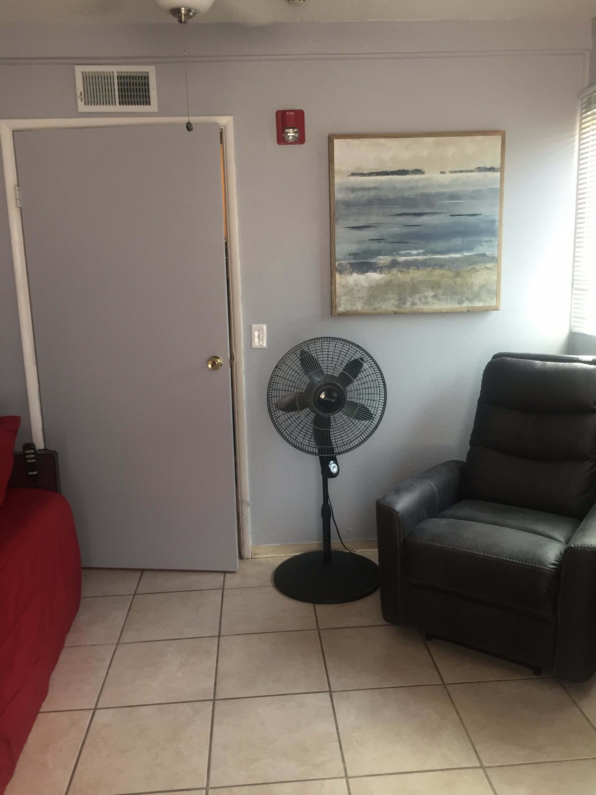 A cozy living space featuring a black armchair, a standing fan, and a seascape painting on the wall.