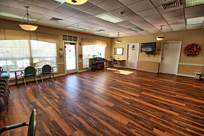 A spacious empty room with wooden flooring, featuring several chairs along the walls, a television mounted on the wall, and ample natural light from large windows.