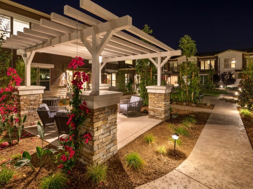 A beautifully lit outdoor seating area under a pergola, surrounded by lush landscaping and pathways at night.