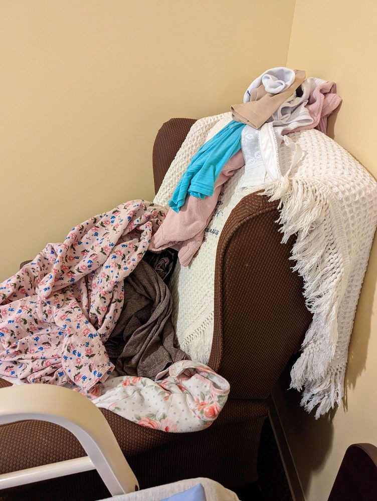 A chair covered with a heap of clothes and a blanket in a cluttered setting.