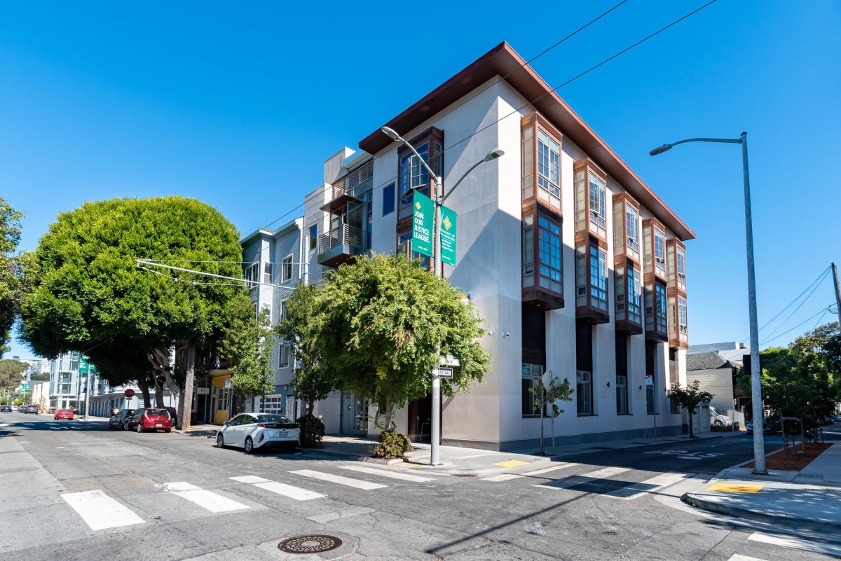A modern apartment building on a sunny street corner with trees nearby.
