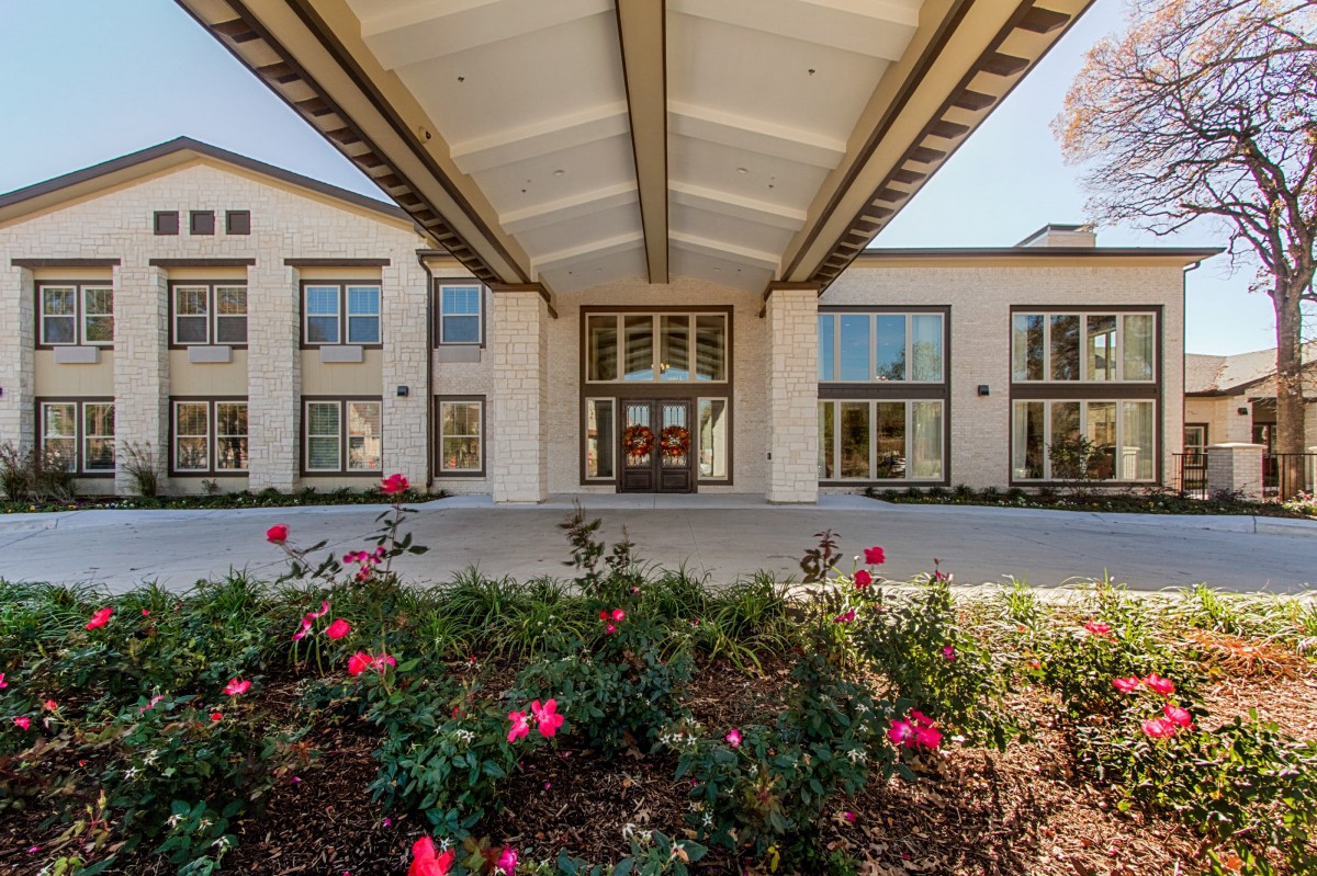 The image shows the front entrance of a modern building with landscaping and vibrant flowers.