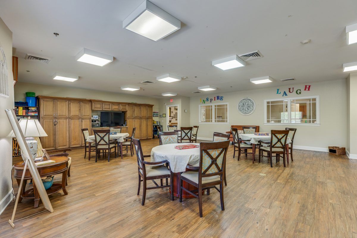 A spacious dining room with multiple tables and chairs, featuring a wooden floor and bright overhead lighting.