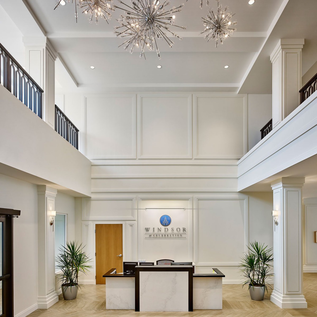 A modern lobby with a reception desk, elegant chandeliers, and decorative plants.