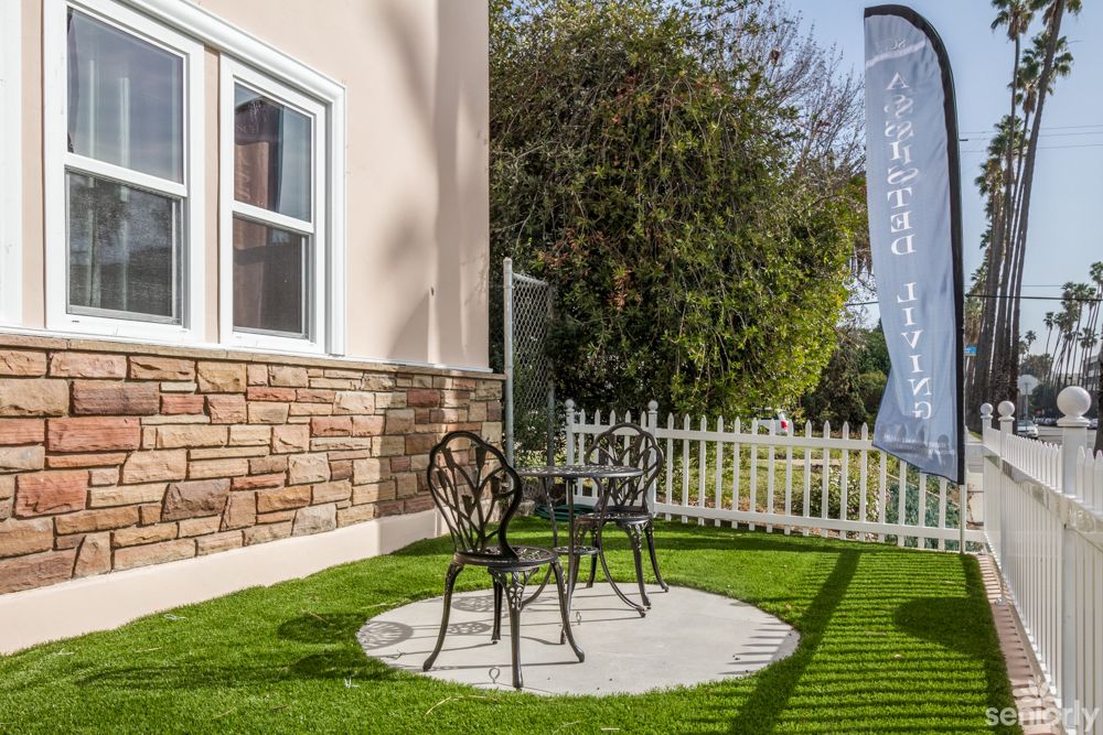 A small patio area with two metal chairs and a table on a round pebble surface, surrounded by a lush green lawn and a white picket fence.