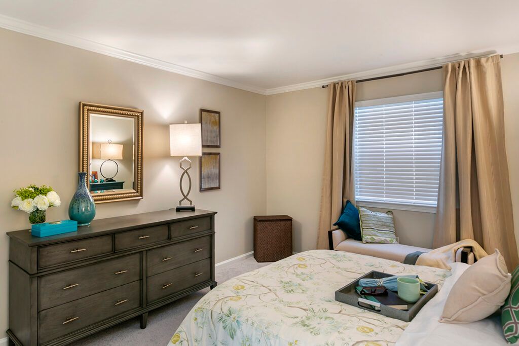 A cozy bedroom with elegant decor, featuring a dresser with a mirror and lamp, along with a bed adorned with cushions and a blanket.
