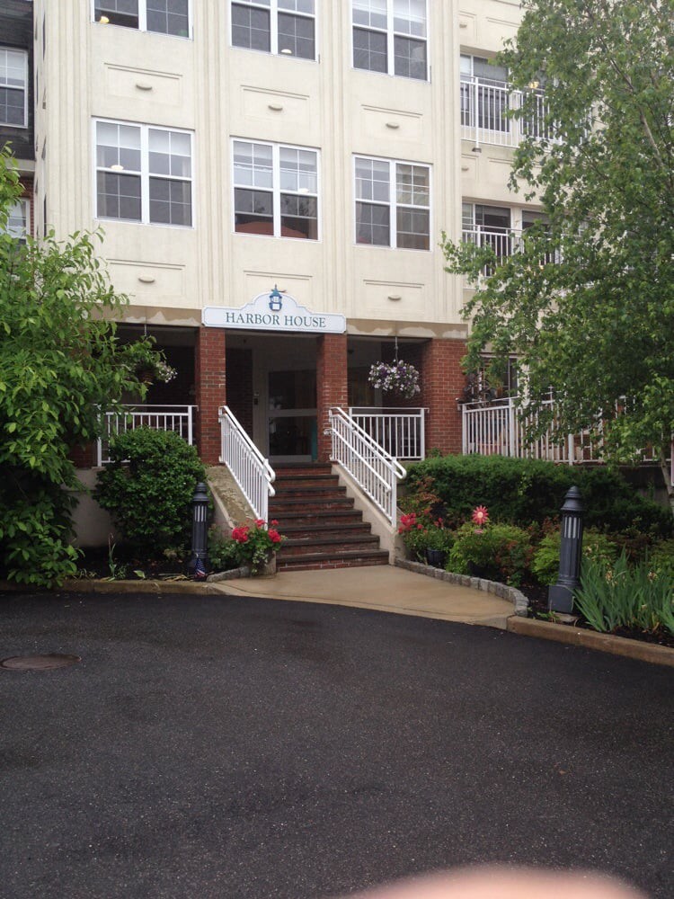 The image shows a building entrance with stairs leading up to the door, surrounded by landscaping with trees and bushes.