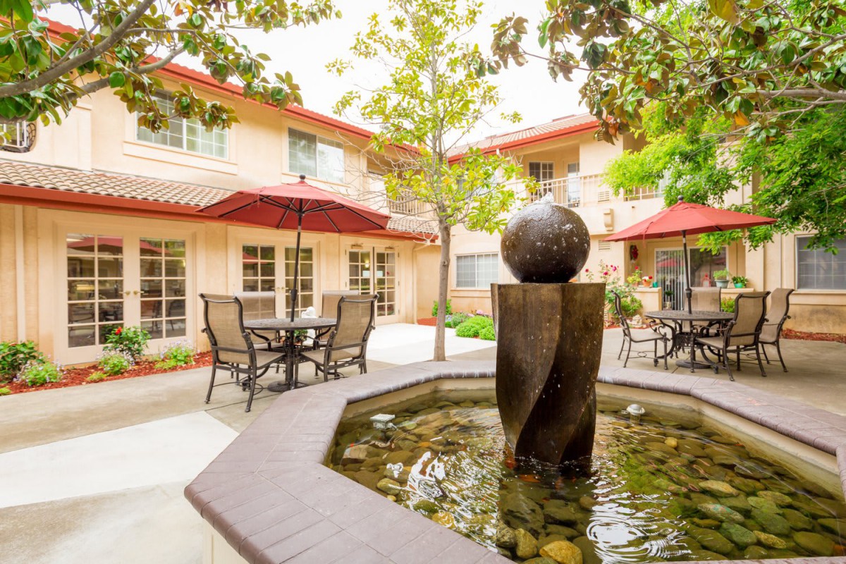 A serene courtyard with a central fountain, outdoor seating, and patio umbrellas surrounded by a two-story building.