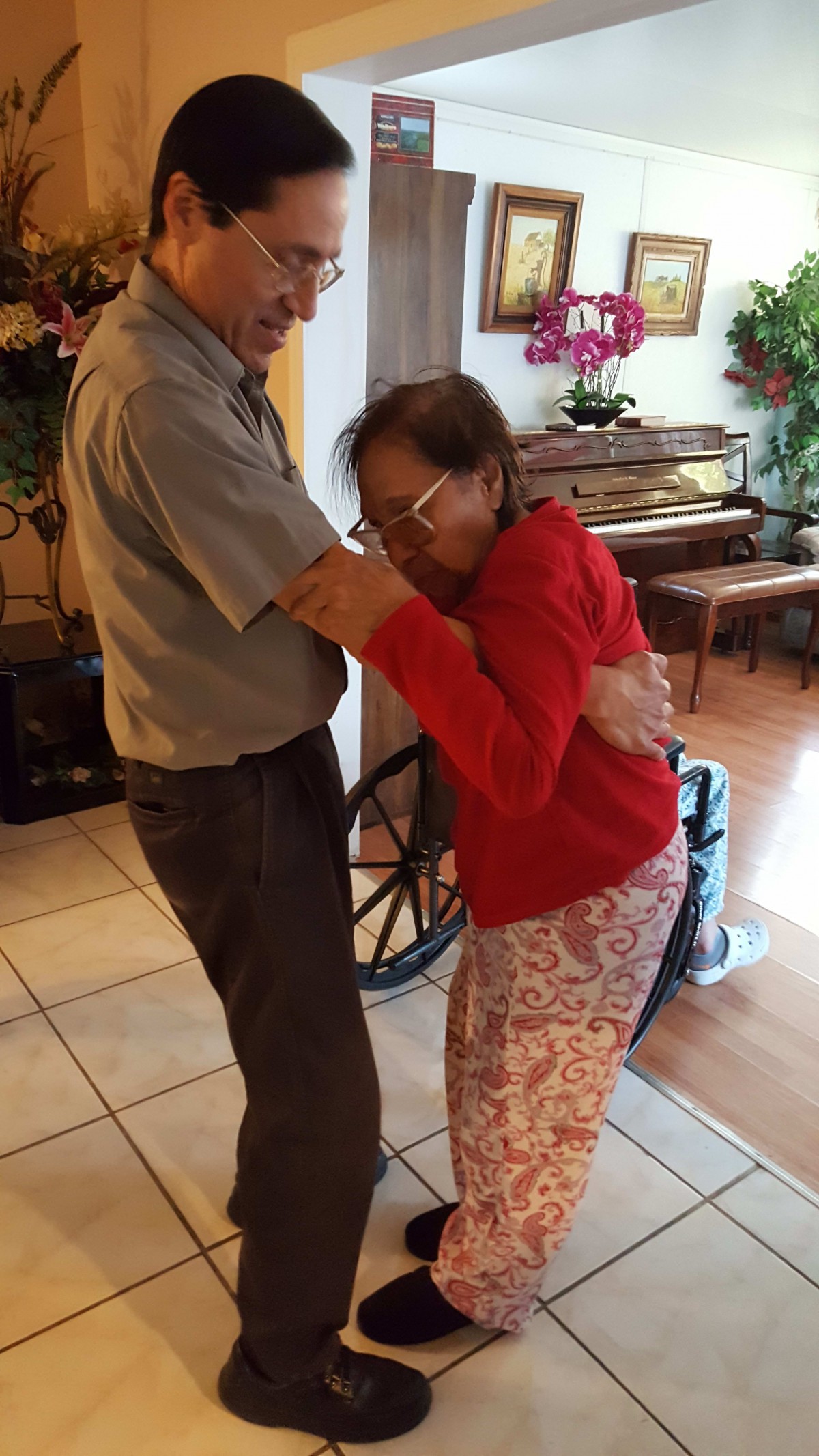 An elderly couple dancing together indoors, with a wheelchair in the background.