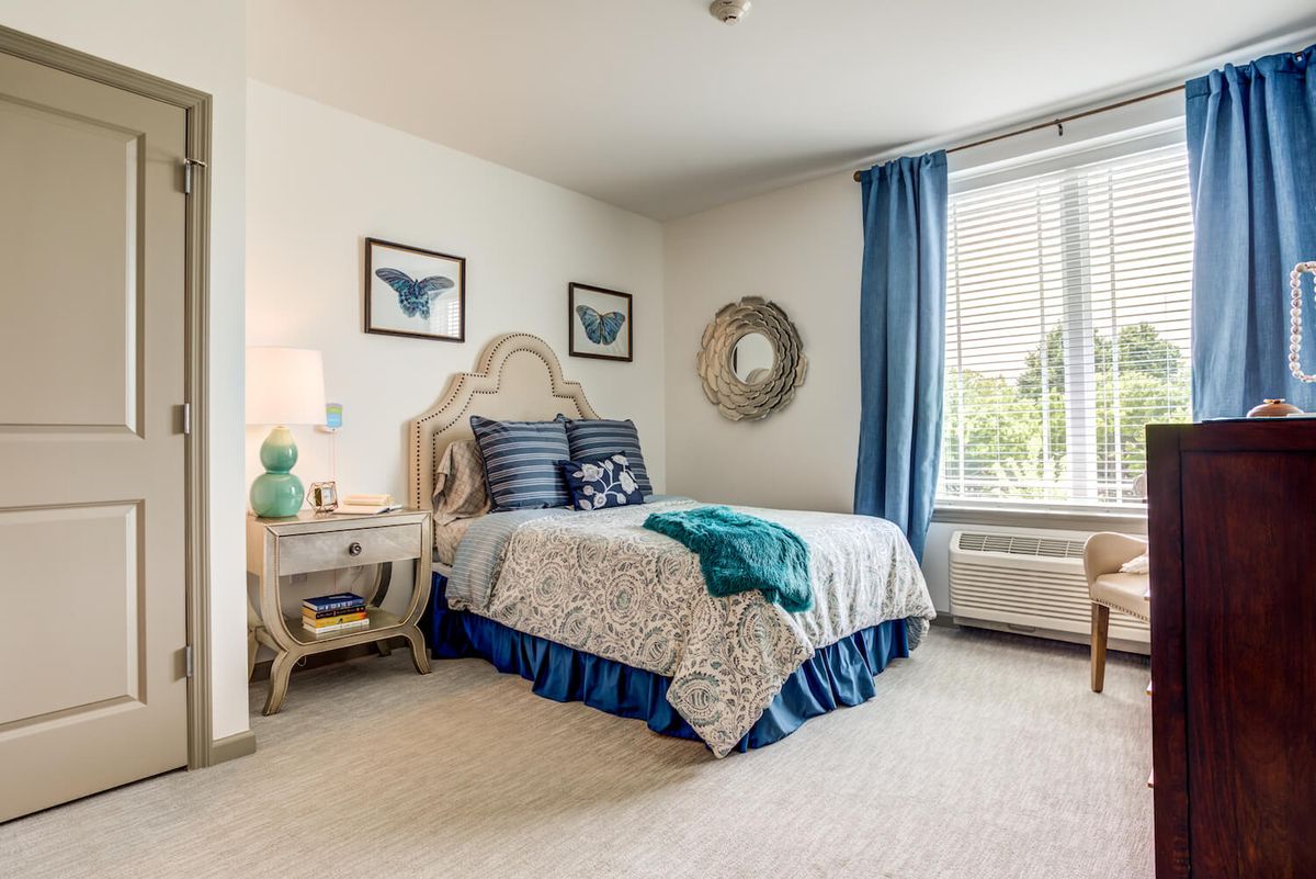 A cozy bedroom featuring a stylish bed with blue accents, wall art, and natural light streaming through large windows.