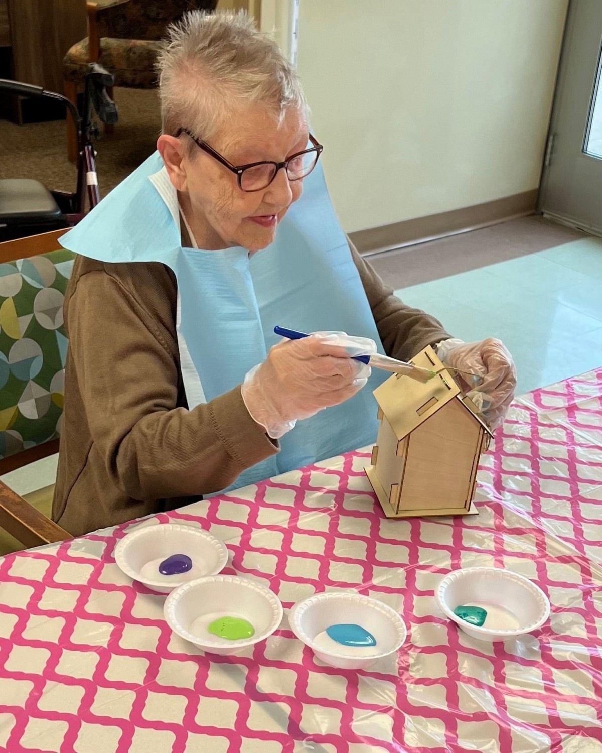 A senior person engages in painting a small wooden structure at a table covered with a red checkered cloth.