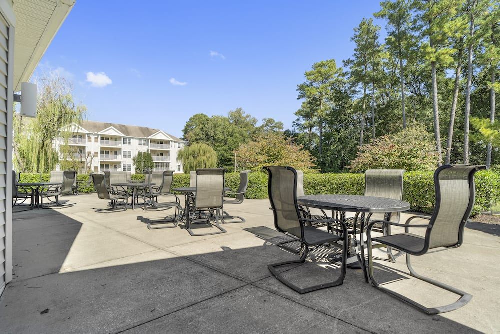 A spacious outdoor patio with tables and chairs on a sunny day, surrounded by greenery and a nearby building.
