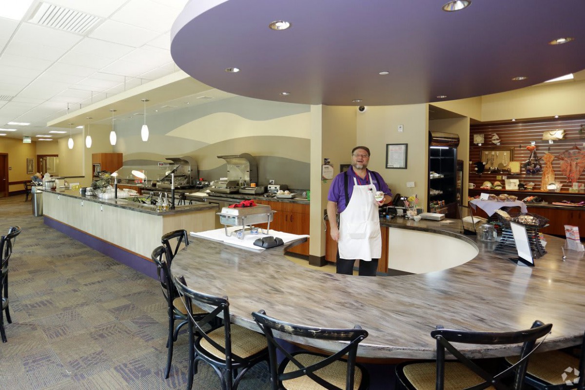 A chef stands behind a curved counter in a modern restaurant, with a buffet setup in the background.