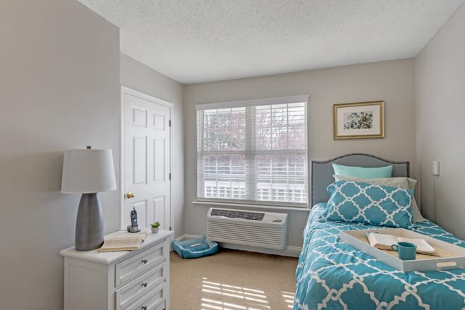 A cozy bedroom with bright natural lighting, featuring a neatly made bed with blue patterned bedding, a bedside table with a lamp, and a window letting in sunlight.