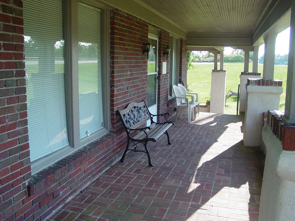 A shaded brick porch with benches and windows overlooking a lawn.