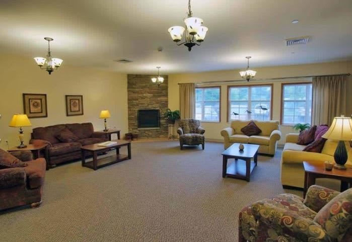 A spacious living room with multiple seating areas, featuring couches and armchairs, a coffee table, a fireplace against the wall, large windows, and chandelier lighting.