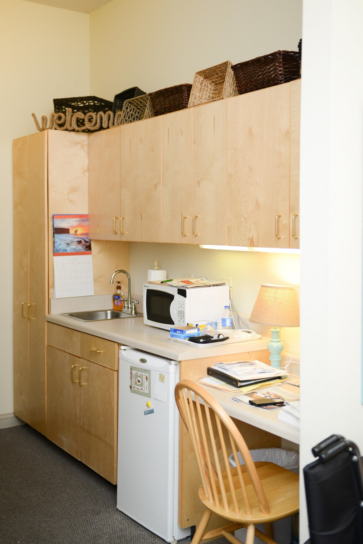 A compact kitchenette with wooden cabinets, a microwave, a small refrigerator, and a workspace with a desk lamp and notepad.