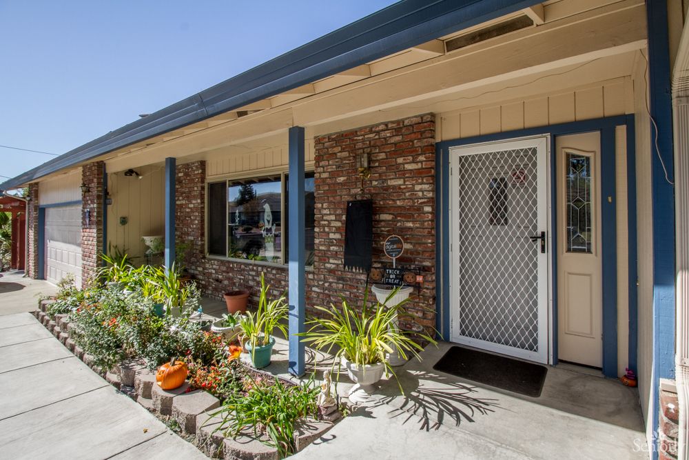 A cozy house exterior with a brick wall, front porch, and a small garden with potted plants.