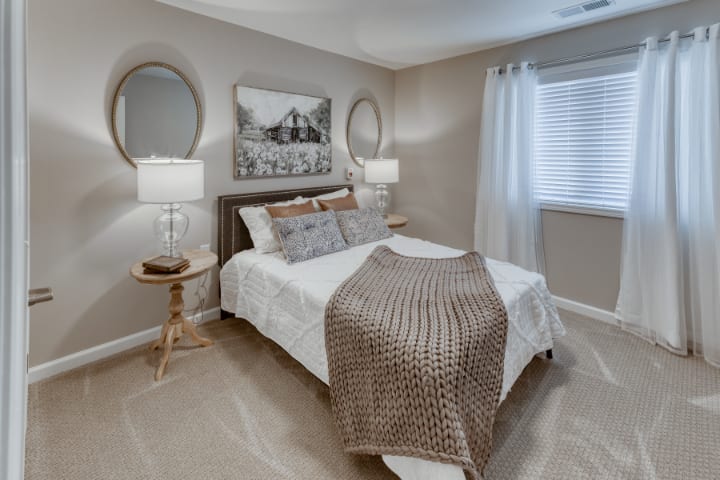 A cozy modern bedroom with neutral tones, featuring a bed with a textured throw blanket, two nightstands with lamps, and decorative mirrors above.