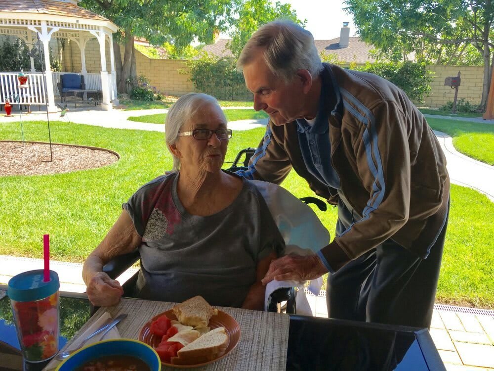 An elderly couple enjoys a conversation outdoors, with a meal laid out on the table.