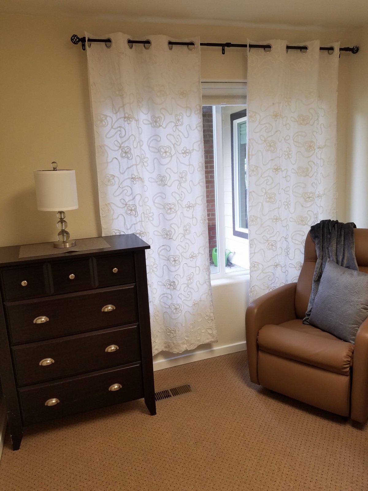 A cozy room featuring a dresser with a lamp, an armchair, and sheer curtains by the window.