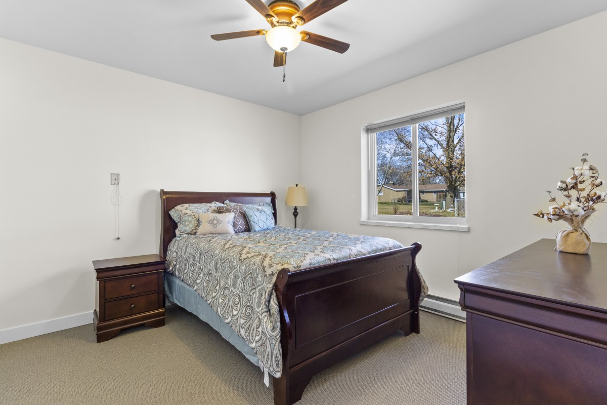 A cozy bedroom featuring a wooden bed with patterned bedding, complemented by a ceiling fan and natural light from the window.