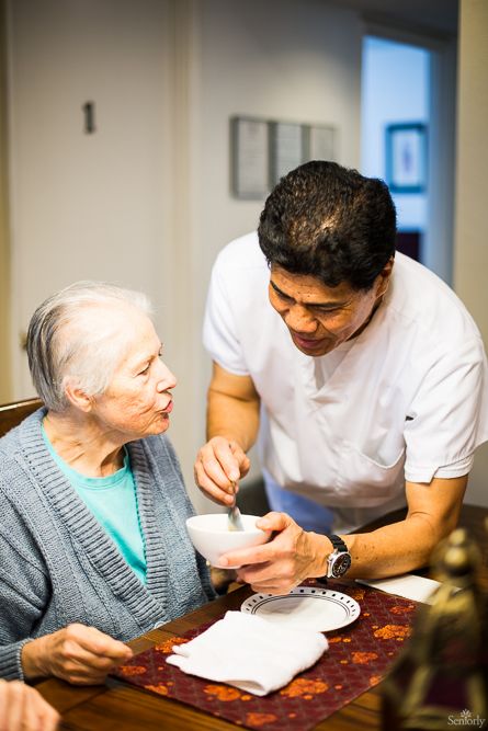 A caregiver assists an elderly woman by feeding her in a warm, home environment.