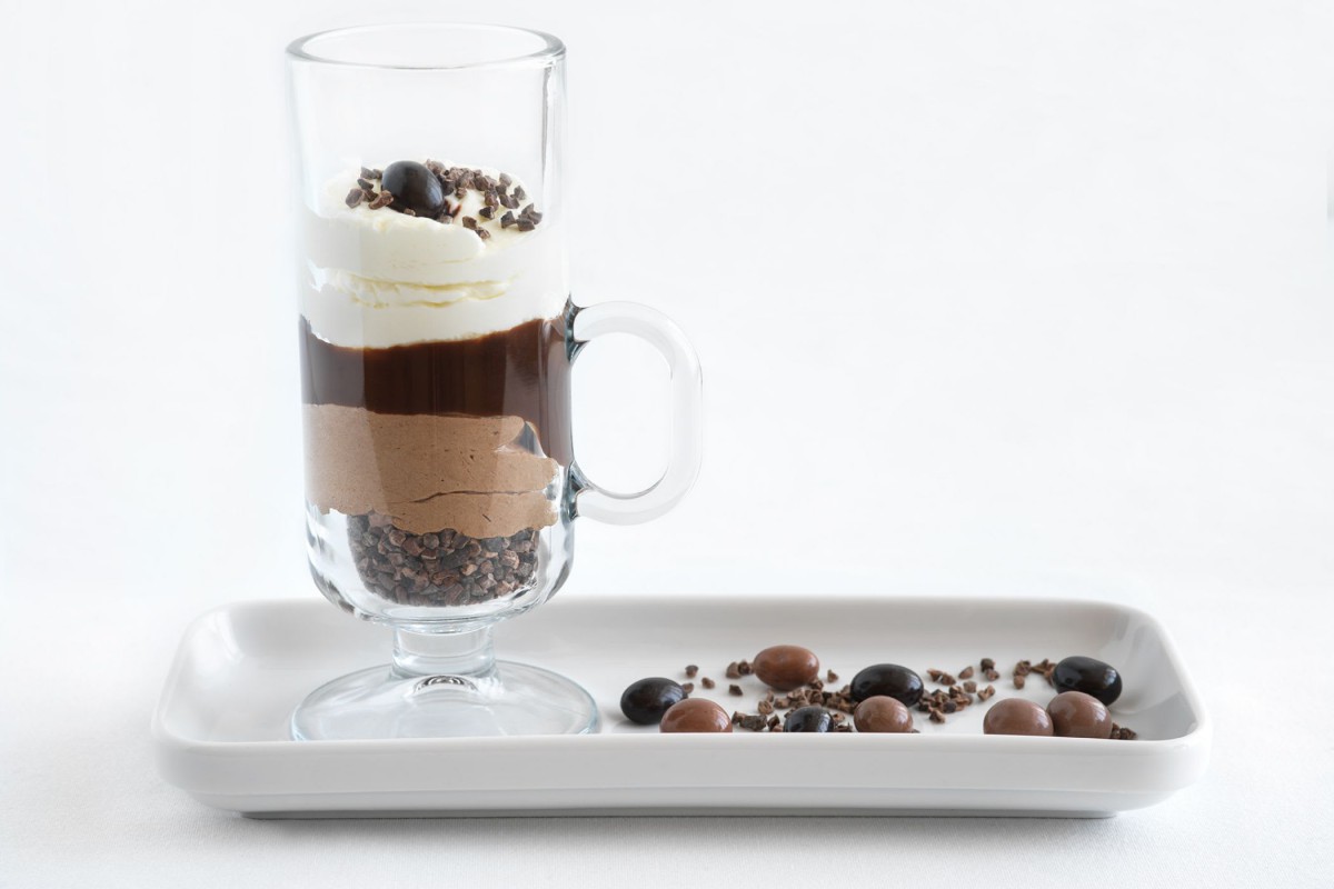 A layered dessert in a glass, topped with whipped cream and chocolate garnish, accompanied by decorative sweets on a tray.